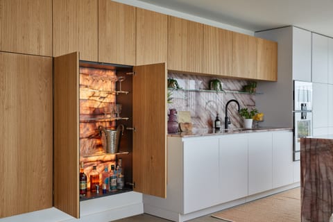 The Clapham drinks cabinet with its routed-oak doors open, revealing a warm backlit interior with glassware and bottles, beside the Cosmopolitan Quartzite run and soft white cabinetry.