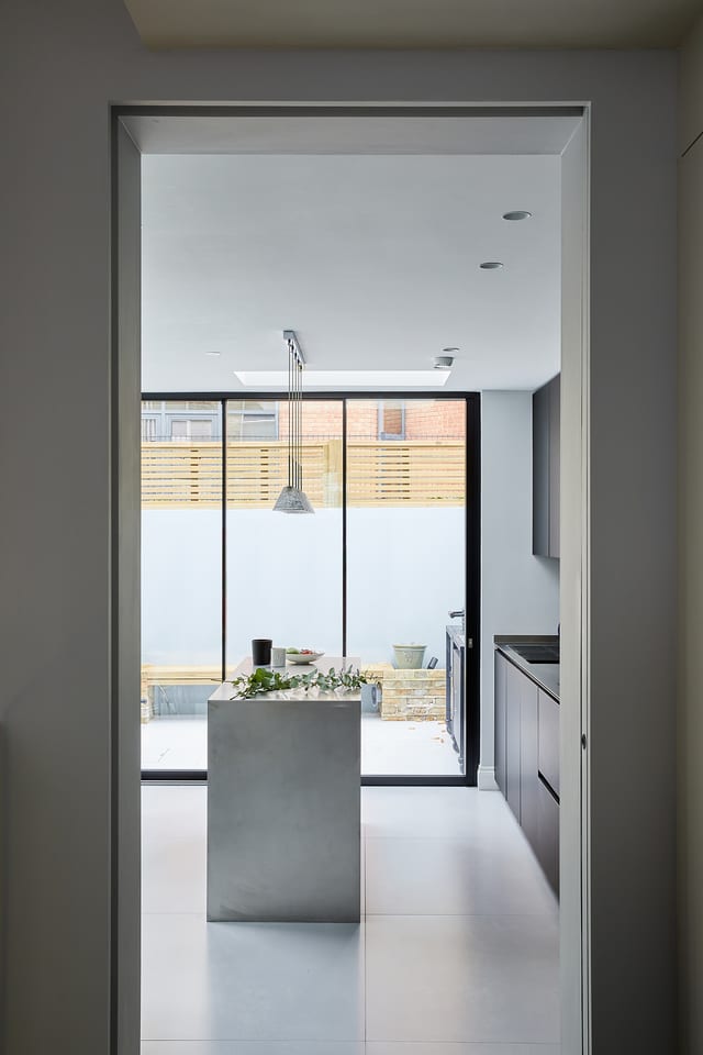 The handmade stainless steel island read as a single monolithic object, framed through the kitchen doorway with the glazed courtyard wall behind.