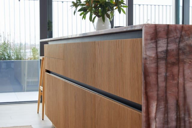 An angled view of the fluted routed-oak island with the Cosmopolitan Quartzite worktop and waterfall edge, the balcony glazing behind.