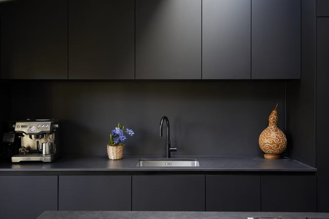 A run of matt dark Eltham cabinetry to the ceiling, with a dark Dekton worktop, an integrated stainless sink and a black tap.