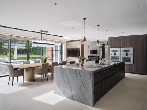 The Keston kitchen seen wide — an angled Platino quartzite island with the cooktop set into it, Leicht Bahia dark timber cabinetry carrying the Gaggenau ovens to the right, and the dining area and patio glazing beyond.