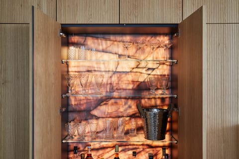 A close view of the backlit drinks cabinet interior — glass shelves of glassware and bottles against a glowing backlit stone back panel.