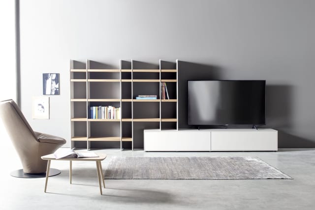 A Sudbrock living-room scene: a low white media unit beside an open timber-and-dark shelving system, with an armchair and a small coffee table.