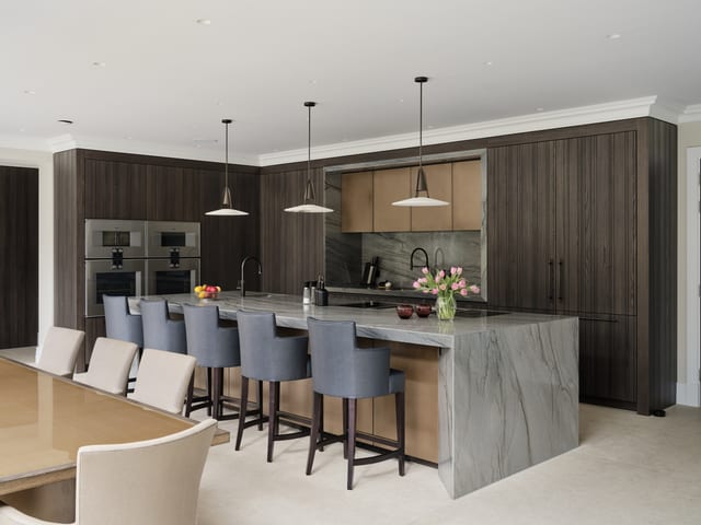 The Keston kitchen from a second angle: the Platino quartzite island with dark upholstered bar stools, Leicht Bahia dark timber cabinetry, a light bronze splashback niche and the Gaggenau ovens to the left.