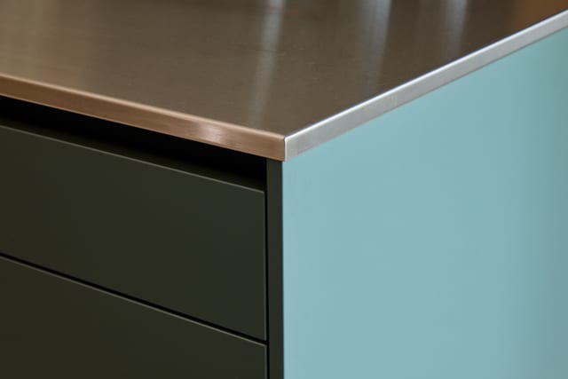 Corner detail of the Hever peninsula: stainless steel worktop edge above; dark Fenix Boreal Green Leicht fronts on one side; a light blue acrylic side panel on the other — the colour accent that gives the project its name.