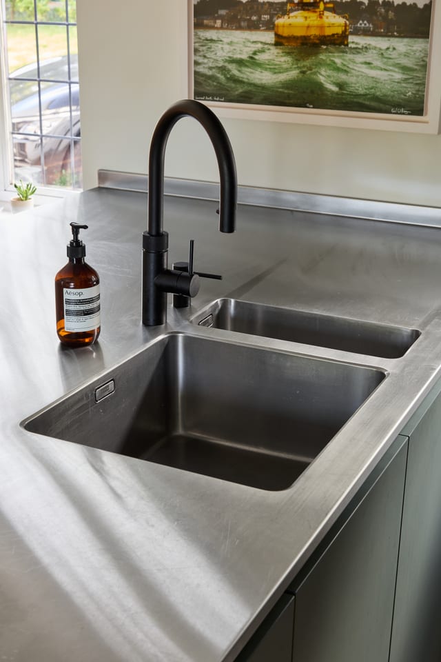 Integrated stainless steel sink and matt black tap on the Hever peninsula, with an Aesop bottle on the counter; the framed buoy print partly visible behind.