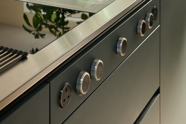 The Bora cooktop on the Hever peninsula, with knurled metal control rings lined up across the dark Fenix Boreal Green drawer fronts and the stainless steel worktop catching the daylight.