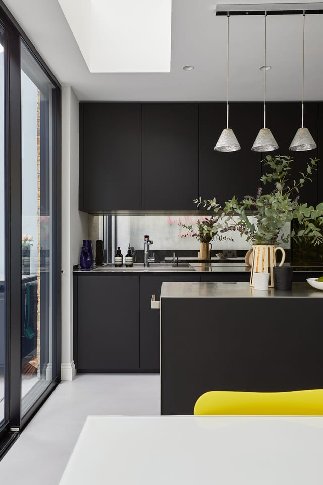 The Hammersmith kitchen: matt black Fenix cabinetry to the ceiling, antique-mirror splashback and stainless worktop, with the stainless island and full-height glazing alongside.