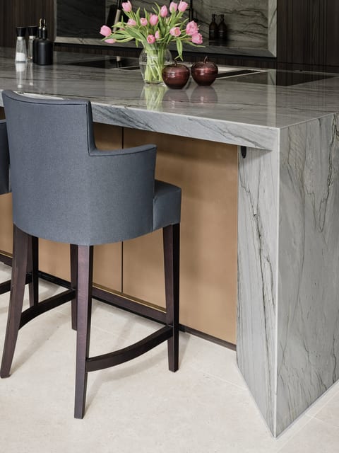 The angled end of the Keston island in Platino quartzite, its grey veining catching the light, with a light bronze panel set into the back and a dark upholstered bar stool alongside.