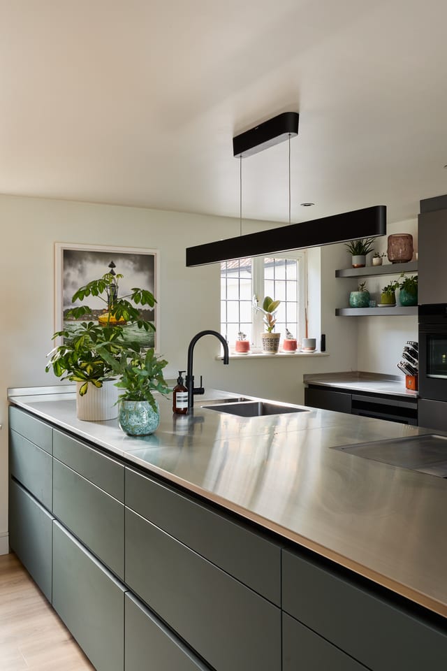 The Hever kitchen peninsula in stainless steel, with potted plants and a black tap on top and dark Fenix Boreal Green Leicht cabinetry below; a framed photograph of an estuary buoy hangs on the wall behind.