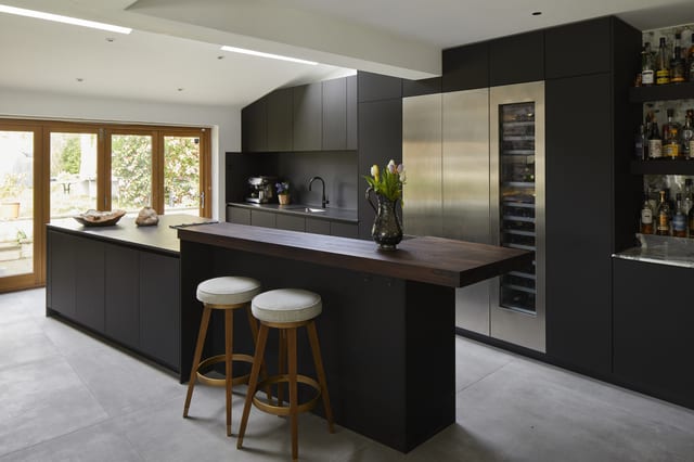 The angled Eltham layout — the dark island with its thick Danish timber top set at an angle to the dark run, the Gaggenau cooling and drinks area beyond, and garden glazing to the side.