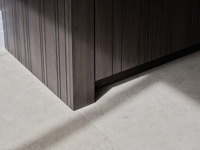 Close detail of the Leicht Bahia routed dark timber, showing the fine vertical grooves where a cabinet meets the stone floor with a recessed shadow gap below.