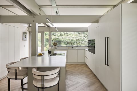 The main Oxted kitchen — white cabinetry and a bronze metallic island with dining chairs, set within the HUF HAUS timber structure, with Caesarstone worktops and garden glazing.
