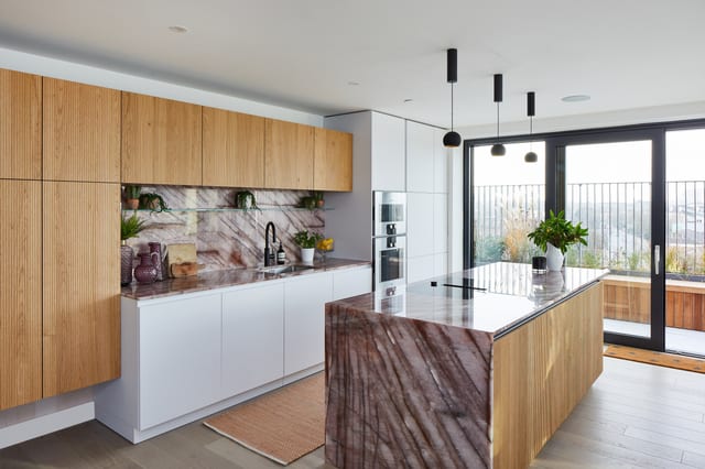 The Clapham kitchen seen wide — soft white Leicht cabinetry and routed oak with a Cosmopolitan Quartzite waterfall island, and glazed doors to a balcony beyond.