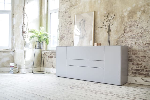 A Sudbrock matt soft-grey sideboard with drawers and doors against a raw plaster and exposed-brick wall, on white-painted floorboards, with a plant on a slim stand and a framed artwork leaning above.