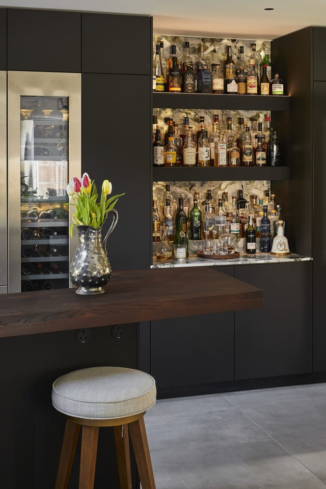 The Eltham drinks area — backlit bottle shelves beside the tall Gaggenau wine cooler in stainless steel, with the Danish timber counter and a bar stool.