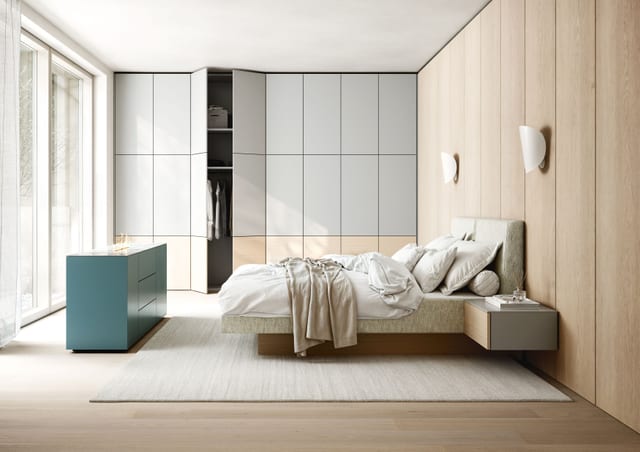 A Sudbrock bedroom furniture scene: a fitted wardrobe wall in soft grey with an open corner section, a muted teal sideboard, an upholstered bed and a floating bedside unit against a timber-panelled wall.