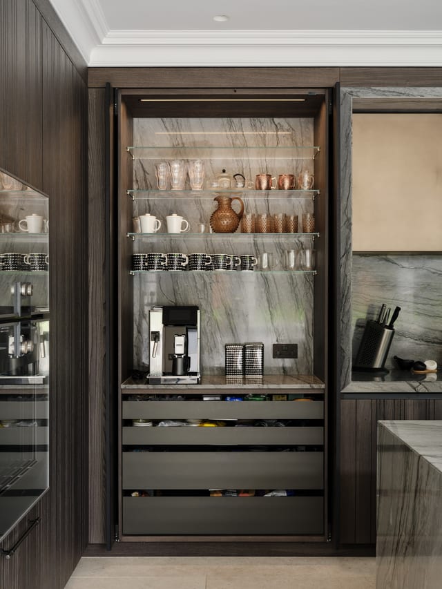 The same Keston cabinetry with the pocket doors slid back to reveal a coffee and breakfast station: a Platino quartzite-lined interior, glass shelves, an espresso machine and open drawers below.