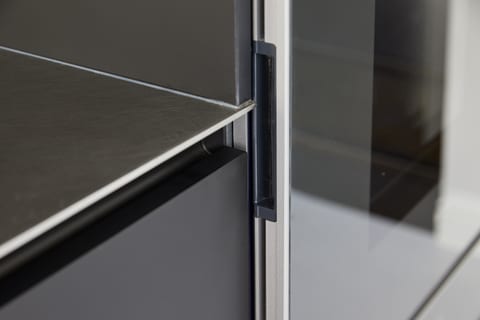 Detail of the stainless steel worktop edge meeting the matt black Fenix cabinetry, with a recessed grip and the antique-mirror splashback above.