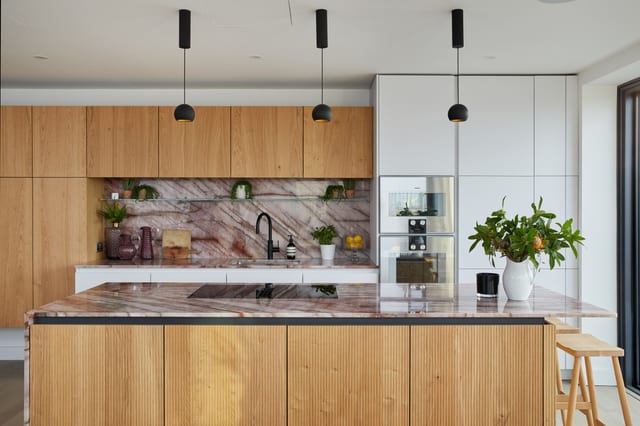 The Clapham kitchen seen wide — a routed-oak island with a Cosmopolitan Quartzite worktop and splashback, soft white Leicht tall cabinetry carrying the Gaggenau appliances, and pendant lights above.